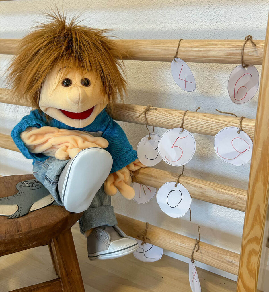 Handpuppe Sam sitzt auf einem Schemel neben einer Sprossenwand.
