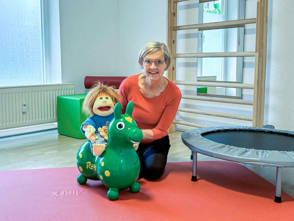 Friederike Schaeff hat Handpuppe Sam auf ein Hüpfpferd aus Gummi gesetzt, neben den beiden ein Fitnesstrampolin, dahinter Sprossenwand und weitere Accessoires für die physiotherapeutische Behandlung.
