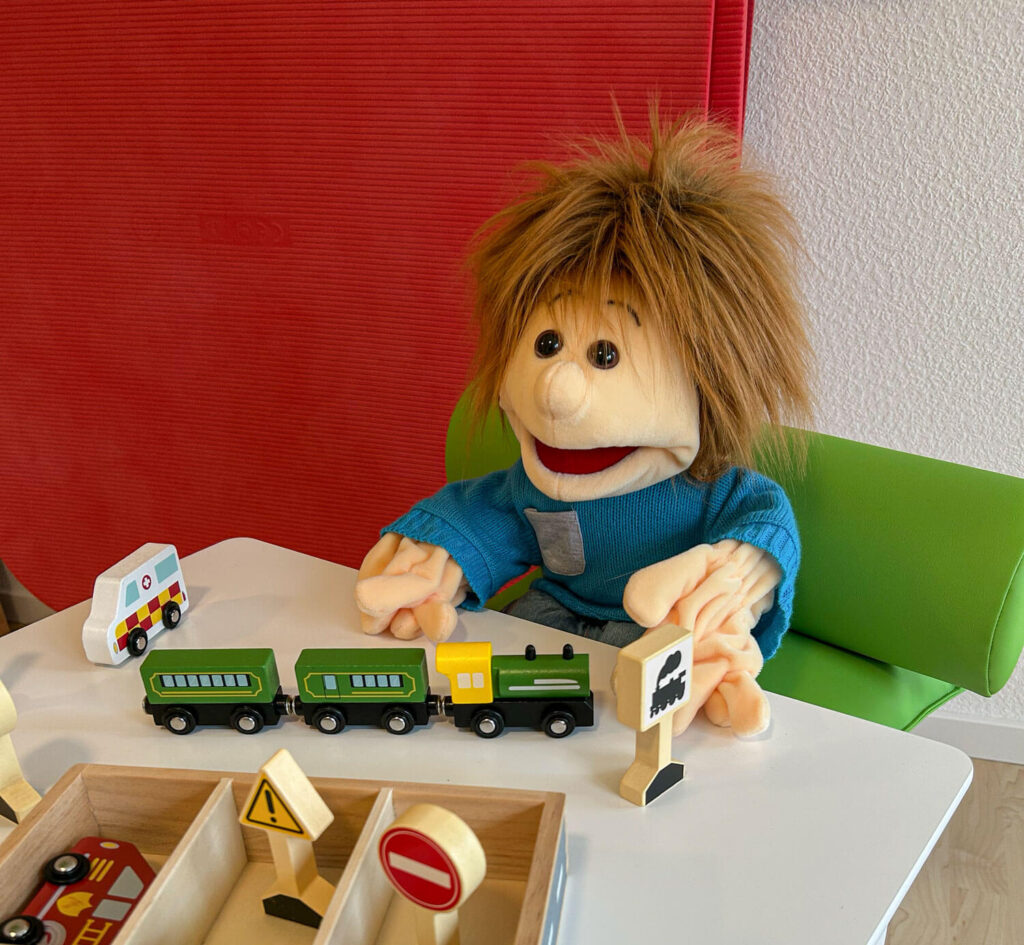 Handpuppe Sam sitzt auf einem Stuhl an einem Kindertisch und hat vor sich eine Spielzeugeisenbahn aus Holz sowie Bahnschilder.