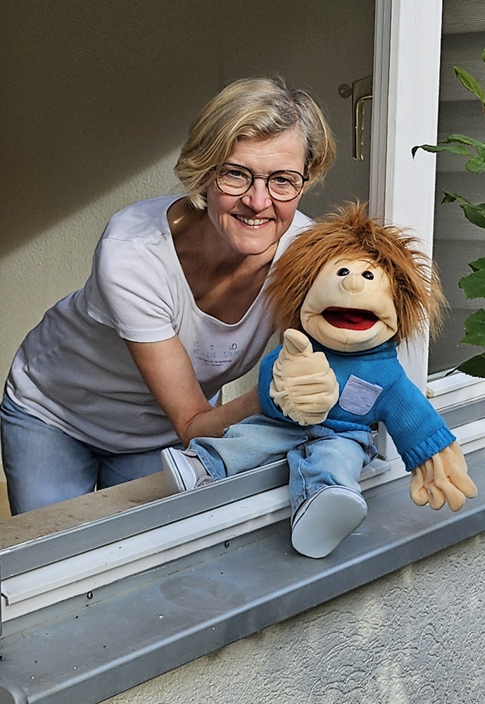 Handpuppe Sam mit "Daumen-hoch"-Geste sitzt im Fesnterrahmen, Friederike Schaeff dahinter, beugt sich in den Fesnterrahmen.