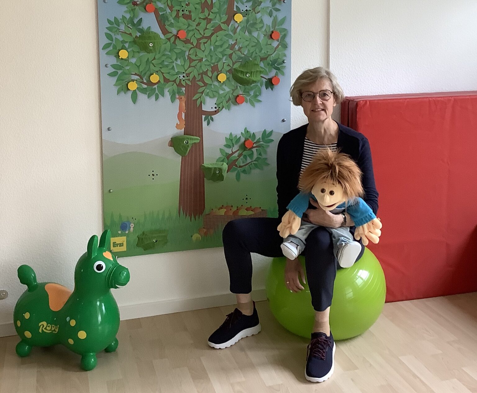 Friederike Schaeff sitzt mit Handpuppe Sam auf einem Therapieball vor der Kletterwand.