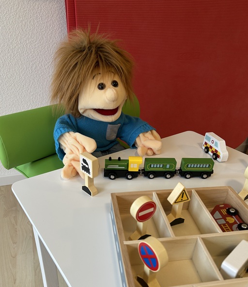 Handpuppe Sam sitzt an einem Kindertisch mit Spielzeug - ein Symbolbild für das Konzept nach Copca, bei dem Kinder dazu angeregt werden, ihr Bewegungspotenzial selbst zu entdecken.