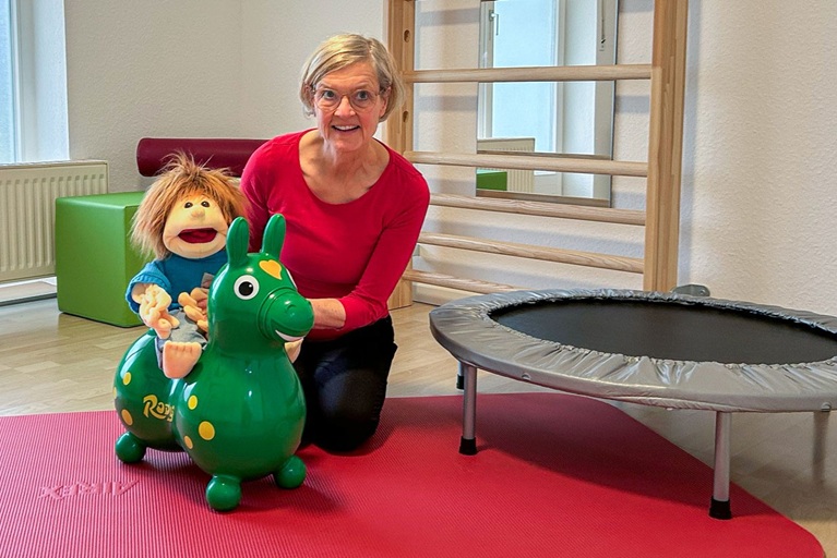 Friederike Schaeff hat Handpuppe Sam auf ein Hüpfpferd aus Gummi gesetzt, neben den beiden ein Fitnesstrampolin, dahinter Sprossenwand und weitere Accessoires für die physiotherapeutische Behandlung.